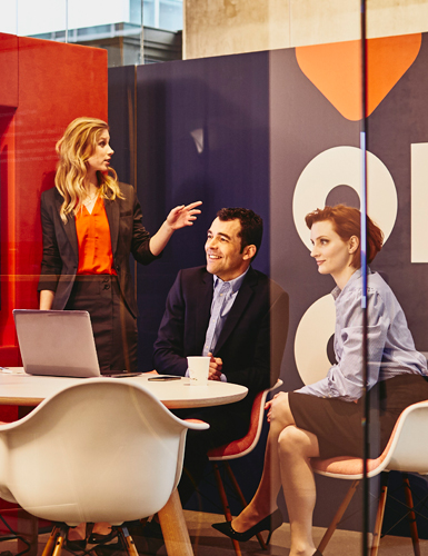 Group of business men and women in a meeting room talking, looking at a screen and a laptop.