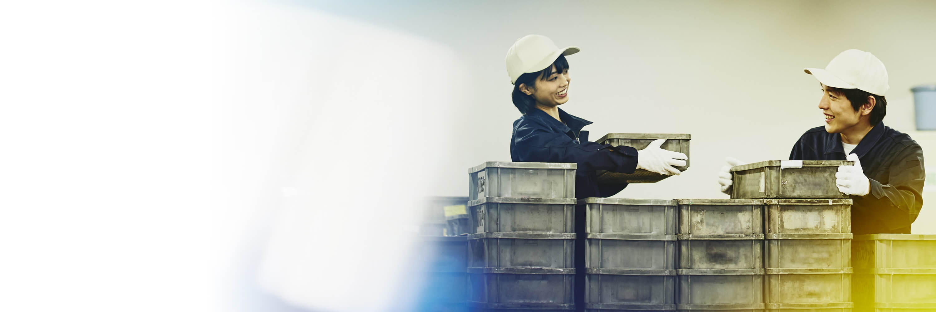 Two colleagues wearing white caps and safety gloves moving boxes in a warehouse.