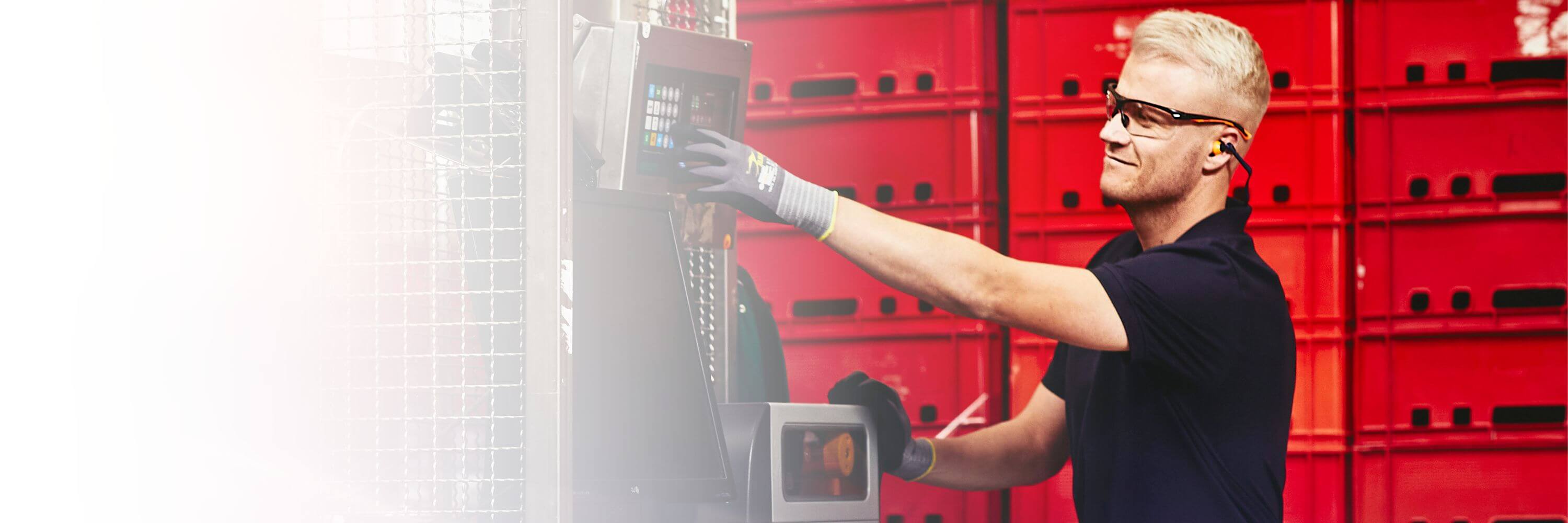 Man wearing safety glasses, gloves and hearing protection, operating a machine in a warehouse.