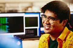 Man smiling while sitting behind his desk, computer screens displaying programming code.