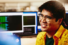 Man smiling while sitting behind his desk, computer screens displaying programming code.