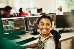 Smiling woman looking away from computer screens displaying financial information.