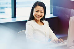 Woman working sitting at her desk in an office. Tall buildings and city in the background.