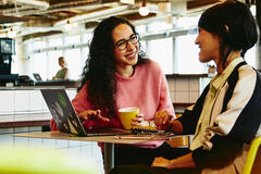 Two woman sitting in restaurant having a converstation, looking at laptop, smiling.