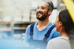 Group of blue-collar workers talking and smiling in a tech environment. Factory/logistics. One male has a groomed beard. The woman has braided hair. Primary color blue. Secondary color cream.