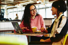 Two woman sitting in restaurant having a converstation, looking at laptop, smiling.