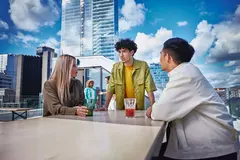 Male in yellow shirt, female and male, standing outside on a balcony at a table, talking