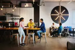 2 male sitting at a table and a female standing at the table and two people sitting at another table