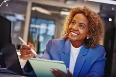 Female with blue blazer holding a touch pen and a tablet, sitting at a desk