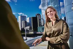Female smiling, standing outside on a balcony talking to a person