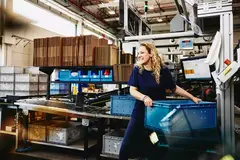 Smiling female standing next to conveyer belt holding a crate
