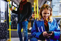 Smiling female sitting in a bus, looking at her phone. Other people in the background.
