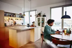 Smiling male sitting at his dining table working on his laptop. Kitchen in the background.
