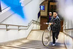 male carrying a bike up the stairs in a train station. Other male walking up the stairs in the background.
