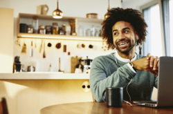man smiling and looking on the side, sitting at a table with a mug and a laptop. Kitchen is in the background.