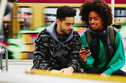 Man and woman looking at phone while sitting on a bench on a train platform.