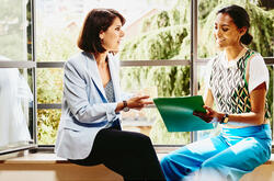 Two woman sitting on a window sill looking at papers.
