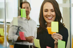 Two business women and a man in an office putting sticky notes on a window.