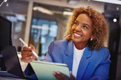 Female with blue blazer holding a touch pen and a tablet, sitting at a desk