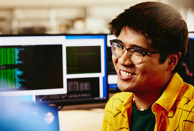 Man smiling while sitting behind his desk, computer screens displaying programming code.