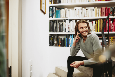 man on the phone smiling and sitting on home stairs with book shelves in the background