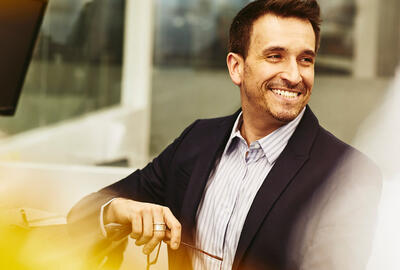 Smiling man in an office holding his glasses while sitting at his desk.