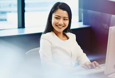 Woman working sitting at her desk in an office. Tall buildings and city in the background.