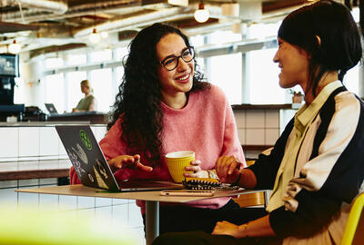 Two woman sitting in restaurant having a converstation, looking at laptop, smiling.