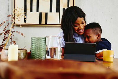 Smiling woman with kid on her lap sitting at a kitchen table with tablet.