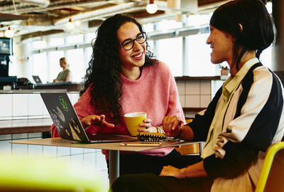 Two woman sitting in restaurant having a converstation, looking at laptop, smiling.