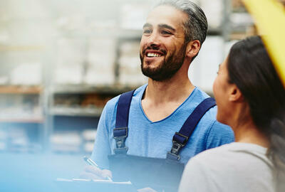 Group of blue-collar workers talking and smiling in a tech environment. Factory/logistics. One male has a groomed beard. The woman has braided hair. Primary color blue. Secondary color cream.