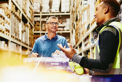 Two man in a warehouse alley with a trolley full of packages.