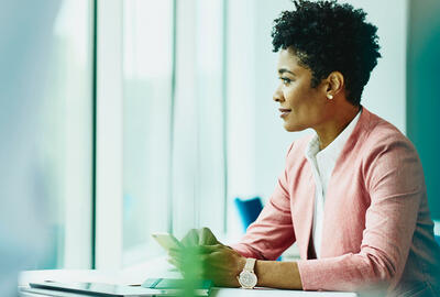 Woman sitting at a table with phone in hand. Tablet and notebook on table. Woman looking out of the window.