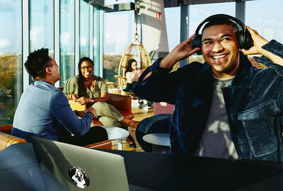 Smiling man with headphones and laptop sitting in a lounge environment.