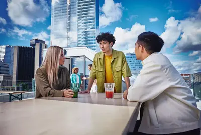 Male in yellow shirt, female and male, standing outside on a balcony at a table, talking