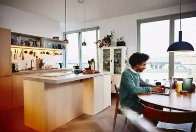 Smiling male sitting at his dining table working on his laptop. Kitchen in the background.
