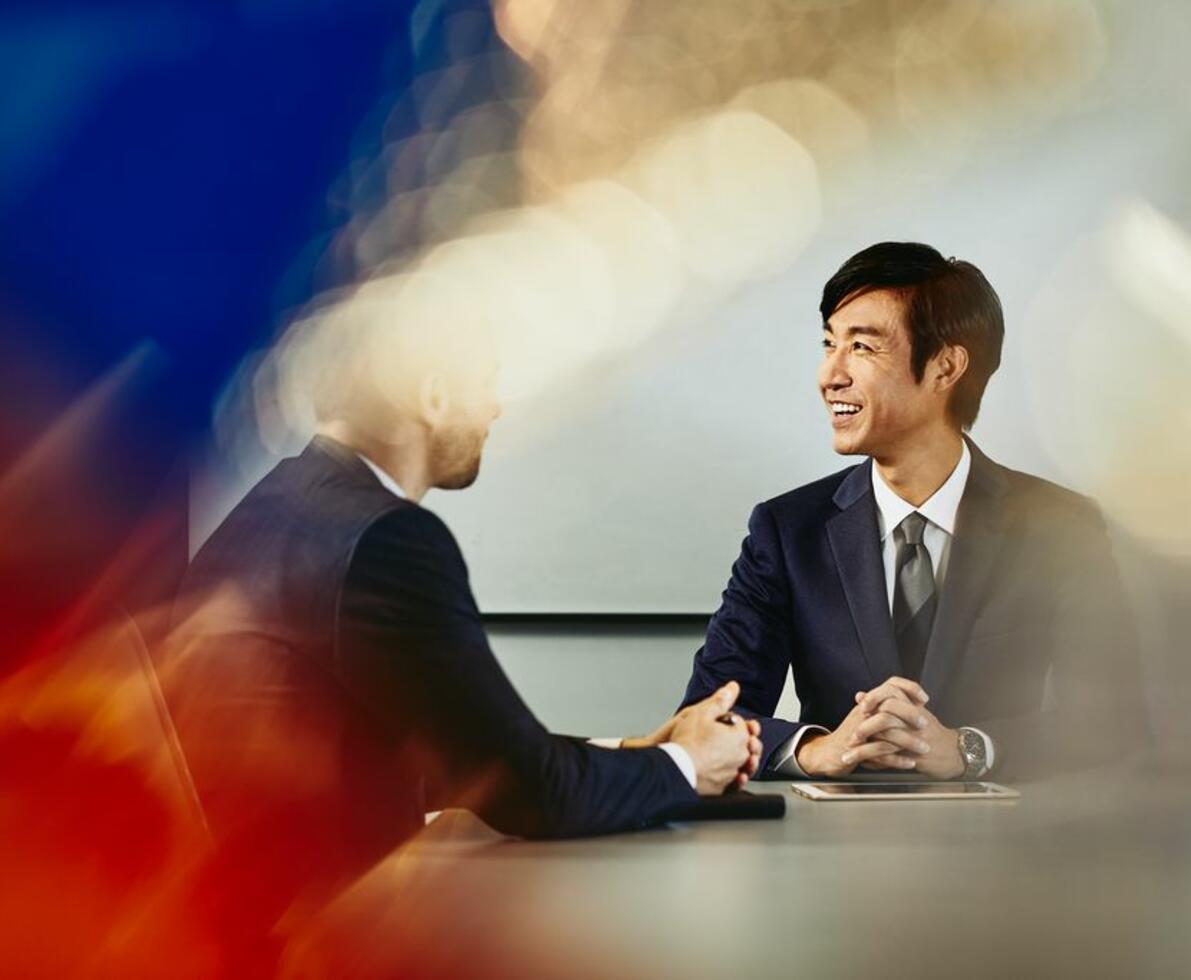 Two business men talking in a meeting, sitting, tablet, whiteboard in the background.