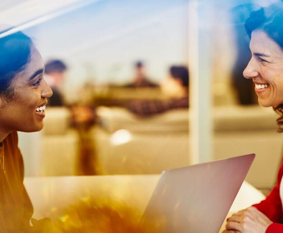 Two smiling women having a conversation in the office with their laptop.