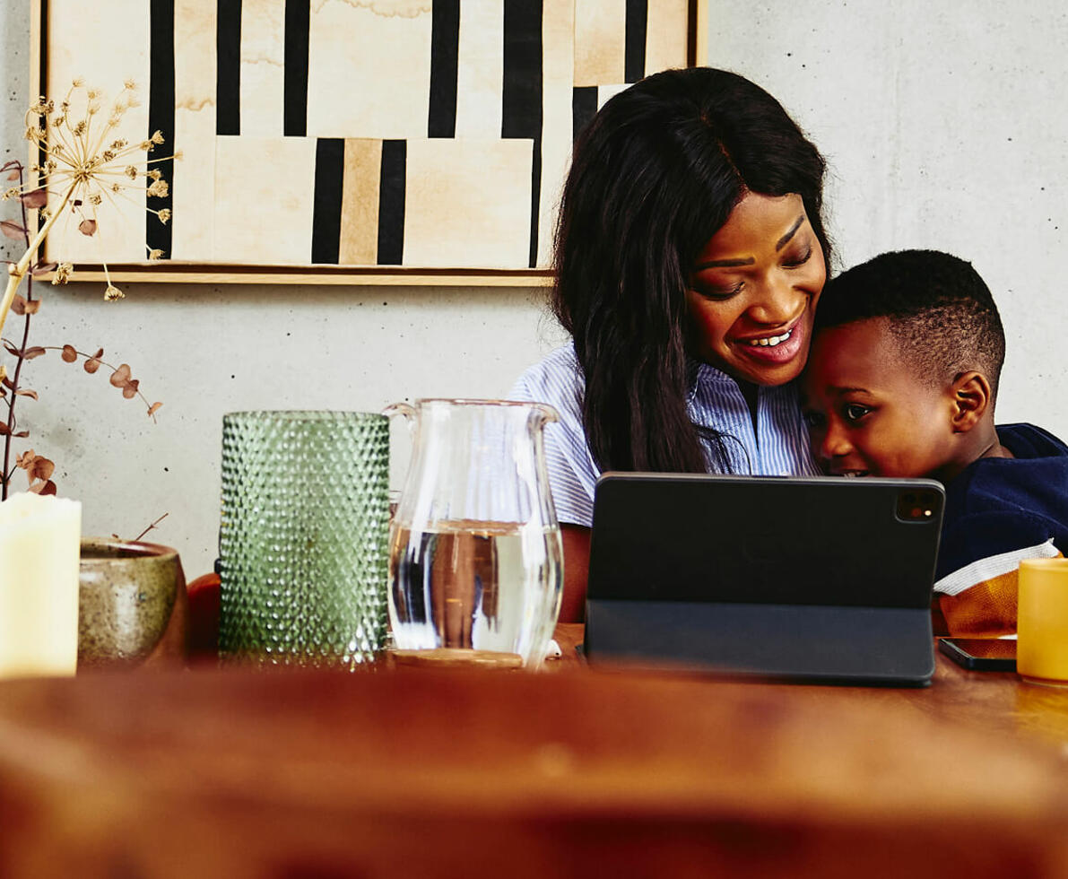 Smiling woman with kid on her lap sitting at a kitchen table with tablet.