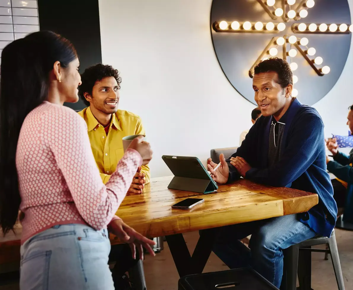 2 male sitting at a table and a female standing at the table with a coffee mug