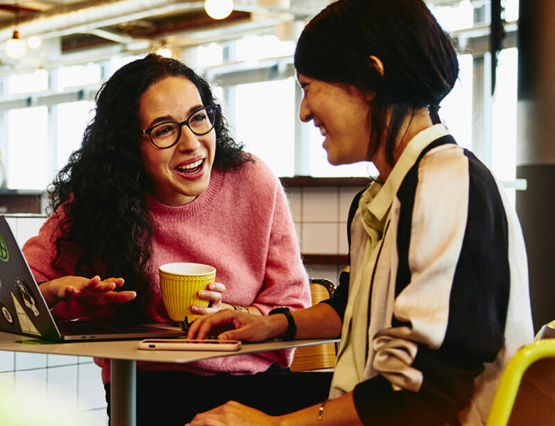 Two woman sitting in restaurant having a converstation, looking at laptop, smiling.