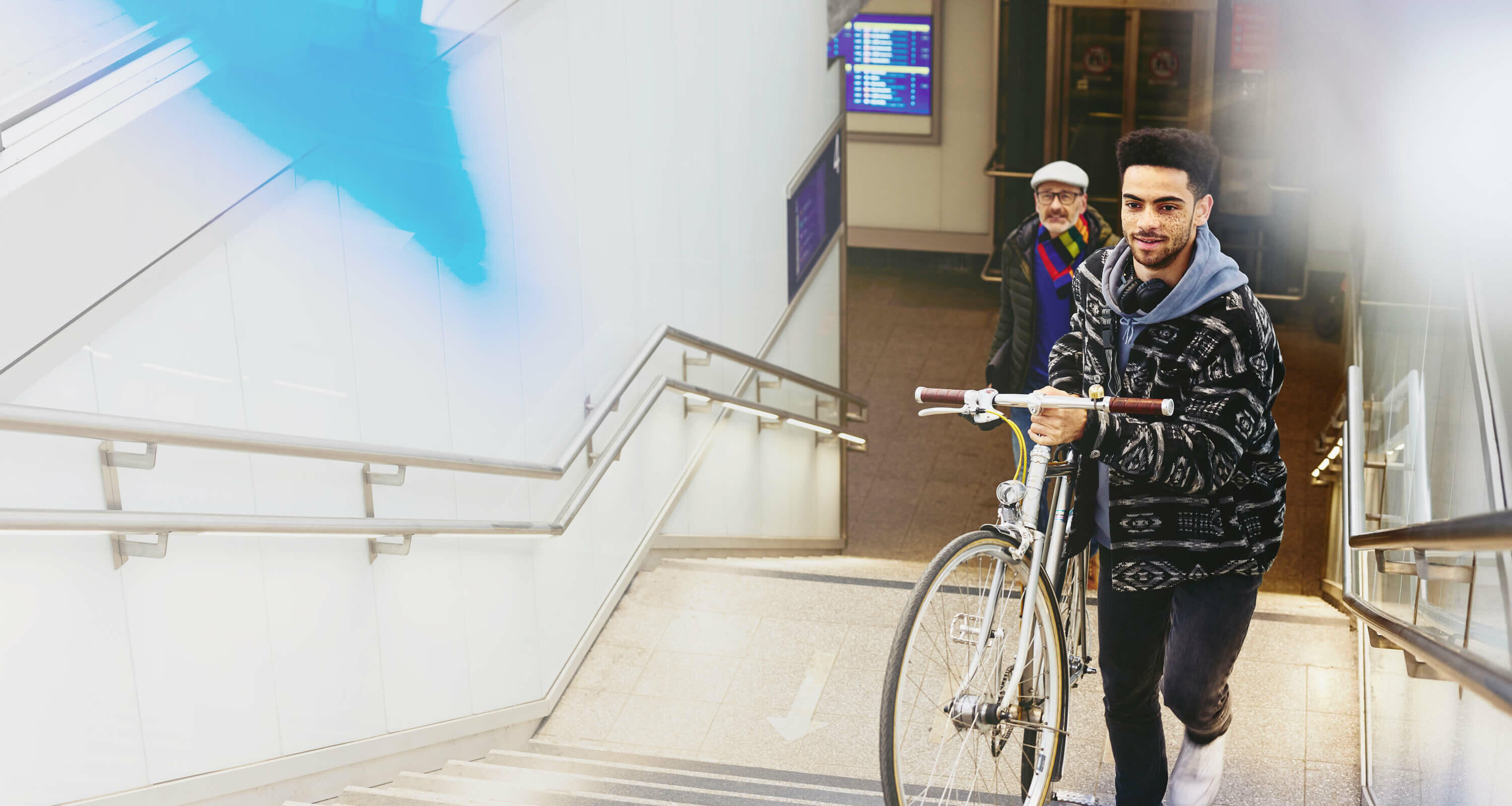 Man carrying a bike up the stairs in a train station. Other man walking up the stairs in the background.