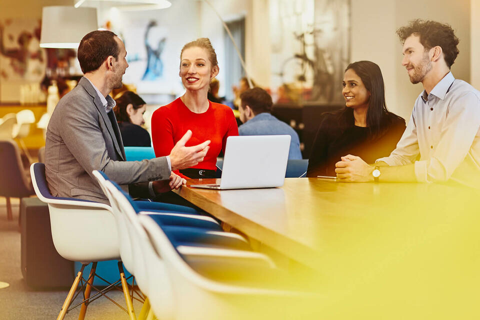 Group of colleagues sitting at a table in a meeting with a laptop, talking and smiling. Office and other people in the background.